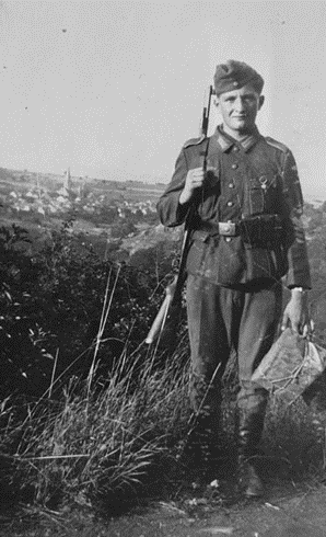 The Cursed Generation. Talking to Wehrmacht Soldiers for the Last Time 4 Black-and-white historical photograph of Jakob, a German soldier in uniform holding a rifle in a rural wartime landscape