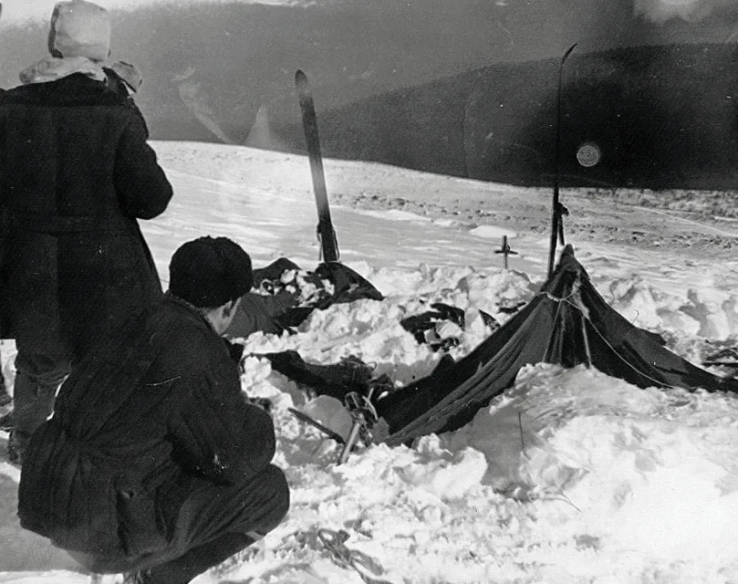 Djatlow-Pass. Schwarz-weiß Fotografie von 1959: Das verlassene und beschädigte Zelt der Djatlow-Gruppe, halb im Schnee vergraben am Hang des Berges Cholat Sjachl. Das zerrissene Zelt liegt schräg im tiefen Schnee, die dunkle Öffnung ist deutlich zu erkennen. Der Himmel ist grau, die Berglandschaft karg und verschneit.